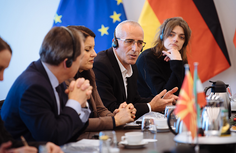 Rund ein Dutzend Personen sitzen an einem Tisch; im Hintergrund sind die europäische und deutsche Flagge zu sehen.